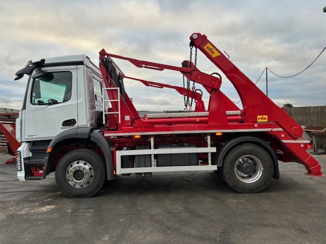 White cab truck equipped with red skip loader arms and hydraulic mechanism parked in an industrial yard.
