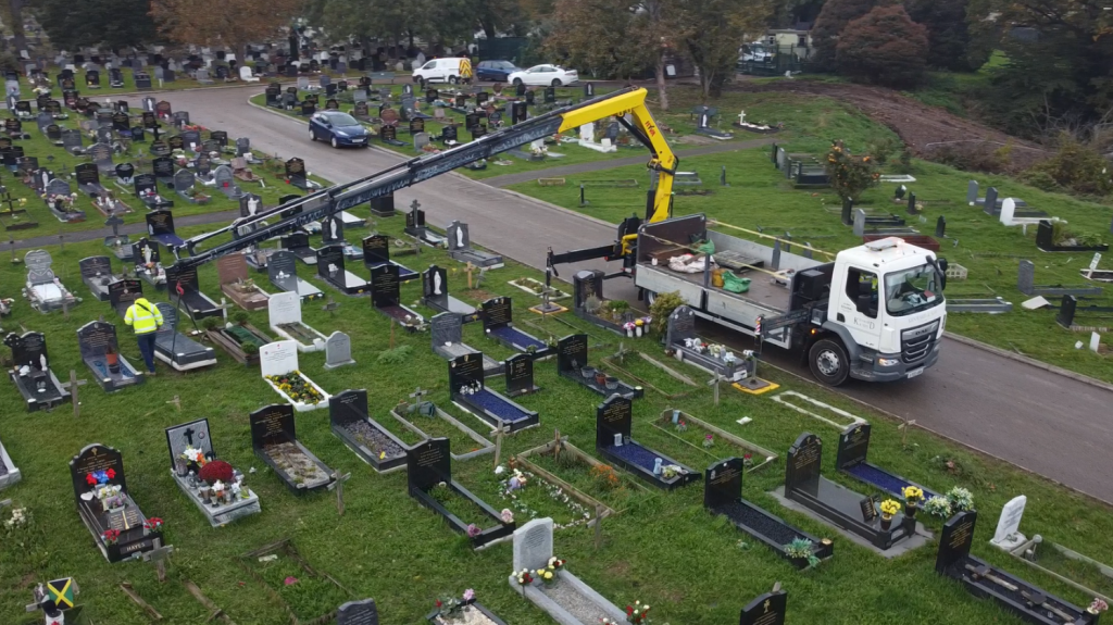 White flatbed lorry with long black telescopic crane and yellow lifting attachment working among headstones in a cemetery.
