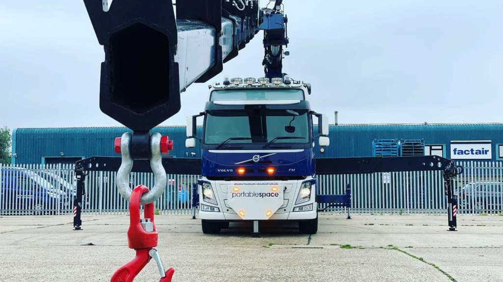 Front view of blue Volvo truck with extended hydraulic crane and red hook in an industrial yard.