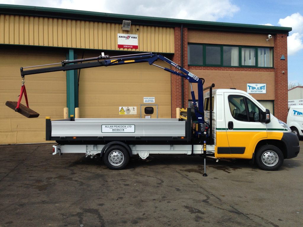 Yellow and white flatbed van with blue hydraulic crane extended lifting a heavy metal block outside a Tip N Lift facility.
