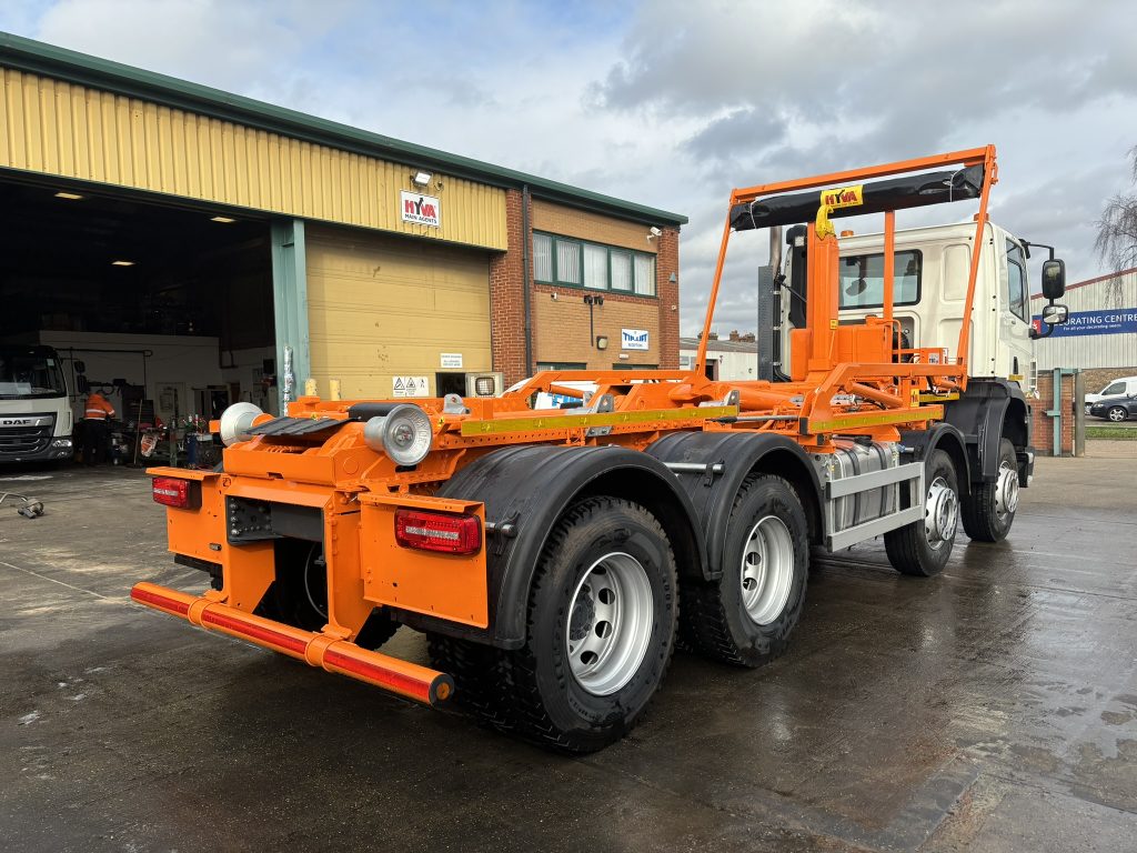 White heavy-duty truck chassis fitted with bright orange hookloader frame parked in a wet industrial yard outside a workshop.
