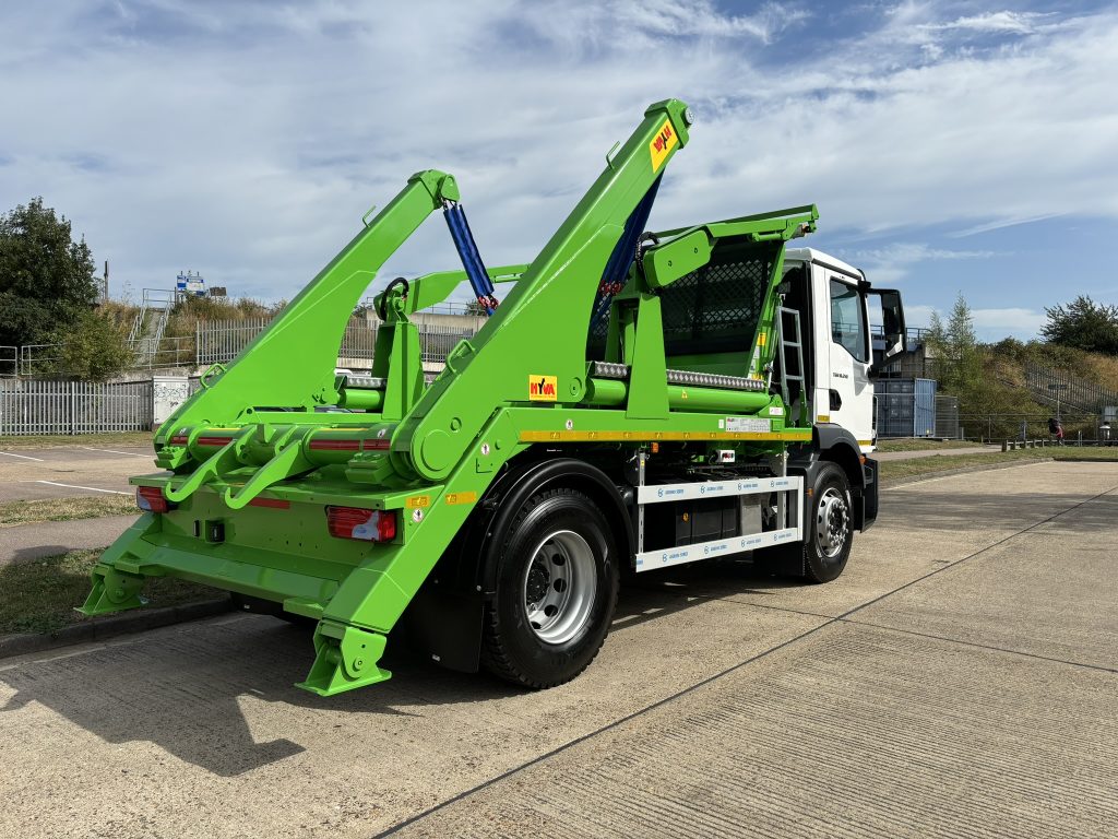White skip loader truck with bright green hydraulic lifting arms parked on a paved surface in an industrial area.
