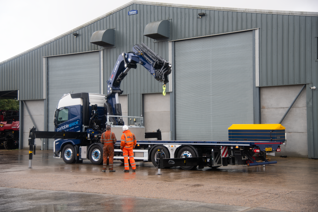 Blue flatbed truck with hydraulic crane extended and two workers in orange high-visibility clothing in an industrial yard.