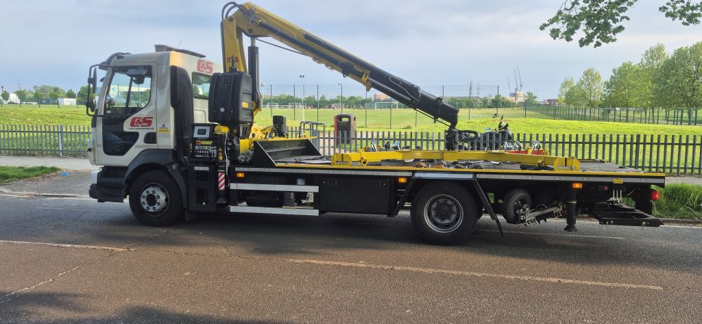 White flatbed truck with C&S branding and folded yellow and black hydraulic crane parked on a wet road near a fenced grassy field.