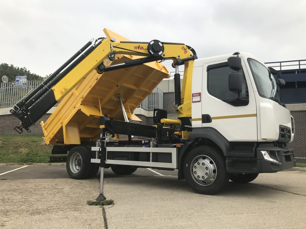 White cab truck with yellow tipping body raised, equipped with a hydraulic crane at an industrial yard.