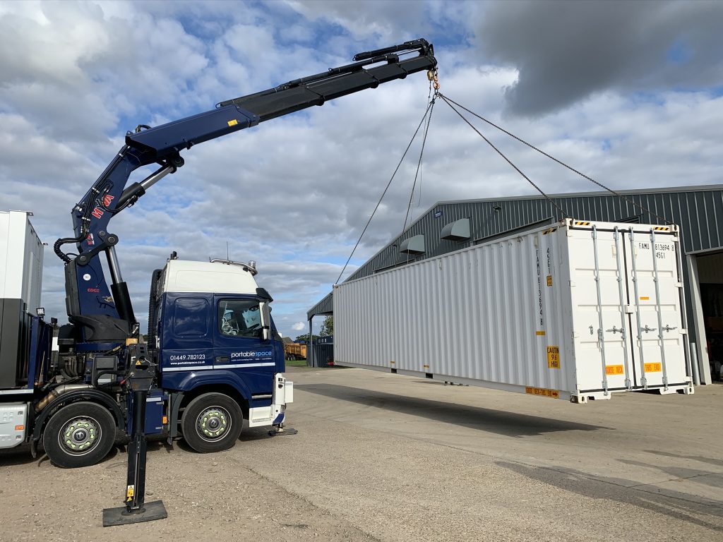 Blue lorry with hydraulic crane lifting a large white shipping container using straps at an industrial site.