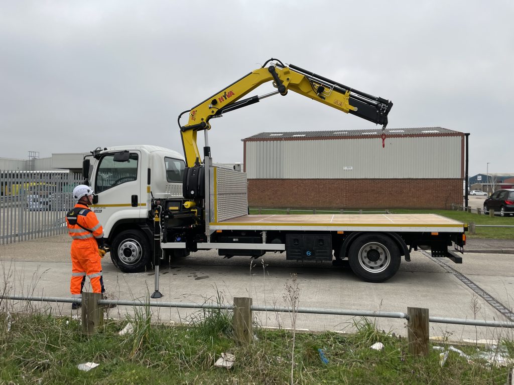 Worker in high-visibility orange clothing operating a yellow truck-mounted crane on a flatbed truck at an industrial site.