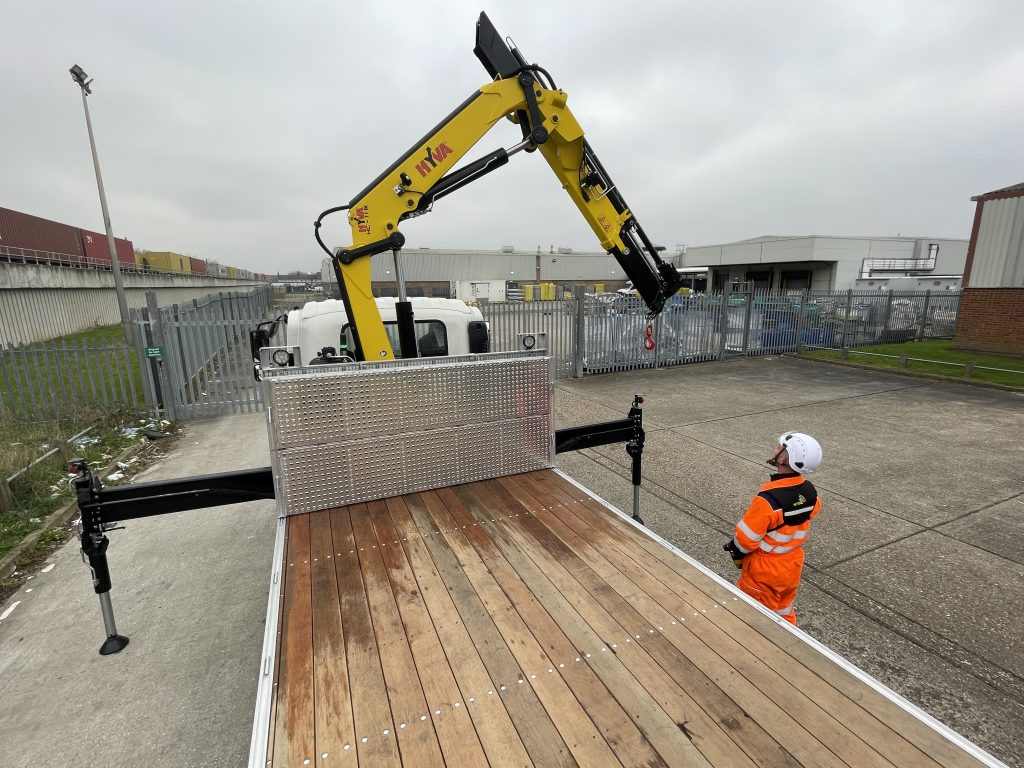 Open flatbed trailer with wooden deck and yellow crane extended, with worker in orange high-visibility gear nearby.