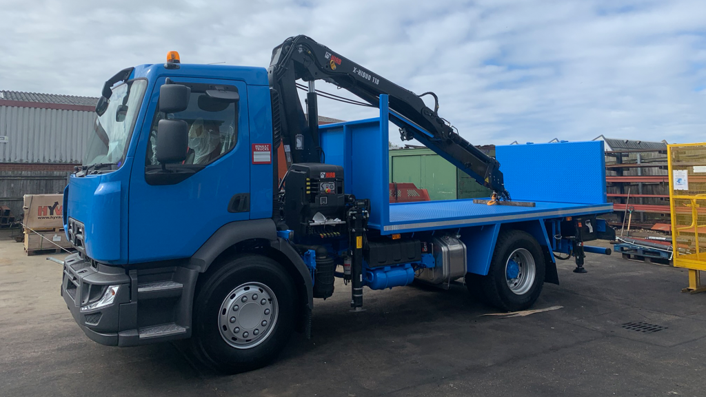 Blue flatbed truck with mounted crane parked in an industrial lot.