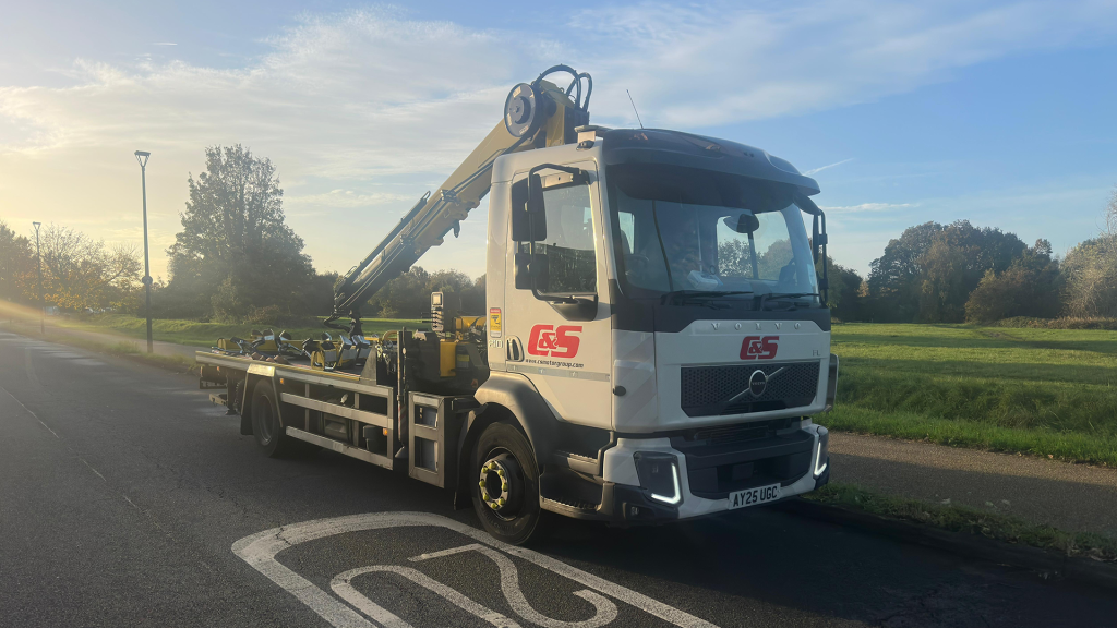 White hook-lift truck with C&S branding and mounted hydraulic crane parked on a road at sunrise with green fields in the background.