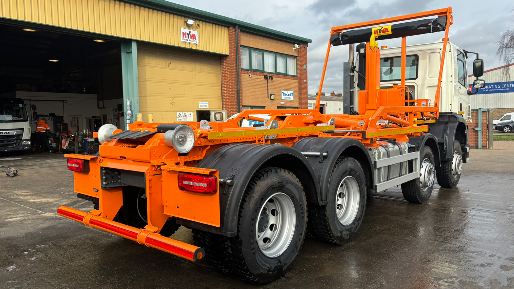 Orange hookloader truck chassis parked outside workshop with heavy-duty hook-lift system.