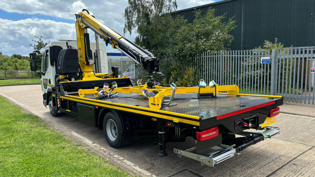 White flatbed truck with a yellow hydraulic crane and tilting platform parked near a metal fence at an industrial site.