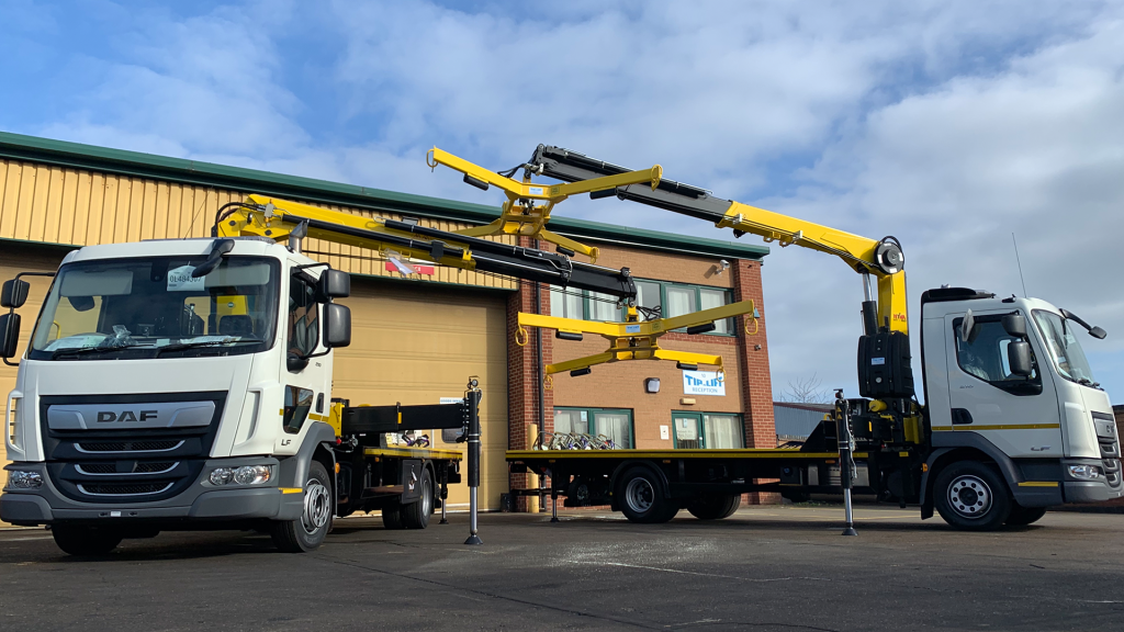 Two white DAF trucks equipped with yellow hydraulic cranes lifting and transporting a modular building panel outside a warehouse.